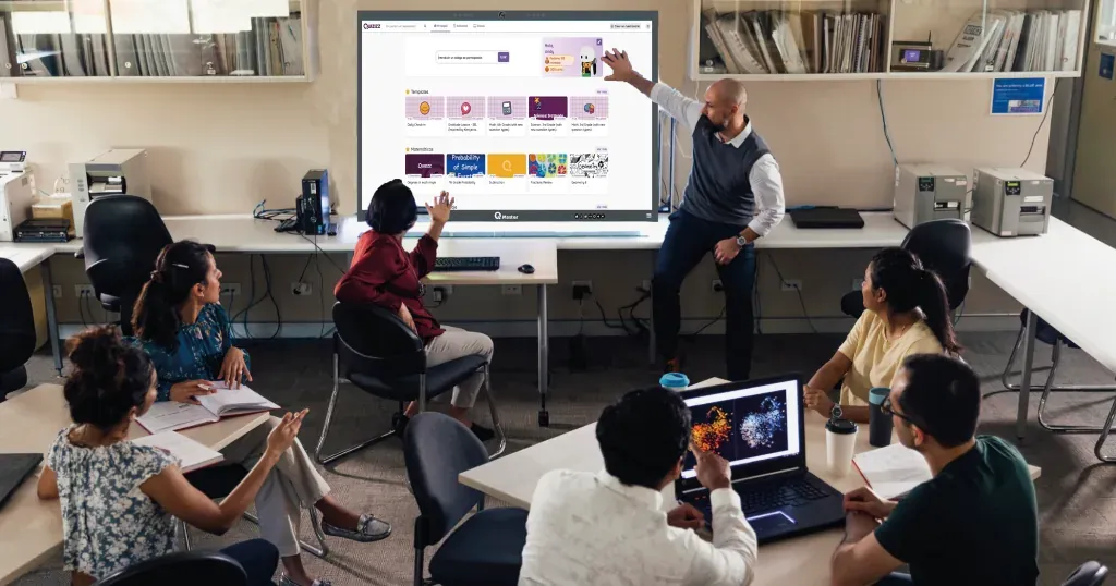 Facilitador señalando contenido en una pantalla interactiva mientras un grupo sigue la sesión desde sus puestos