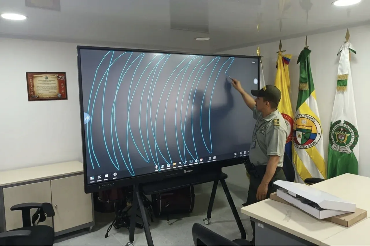 Un uniformado traza sobre la pantalla interactiva durante un ejercicio en el aula de formación, junto a las banderas institucionales.