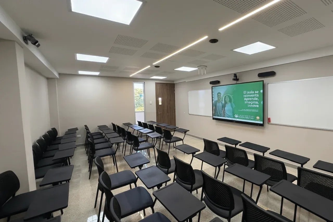 Salón con la pantalla interactiva encendida junto al tablero y el mobiliario dispuesto para la clase presencial.