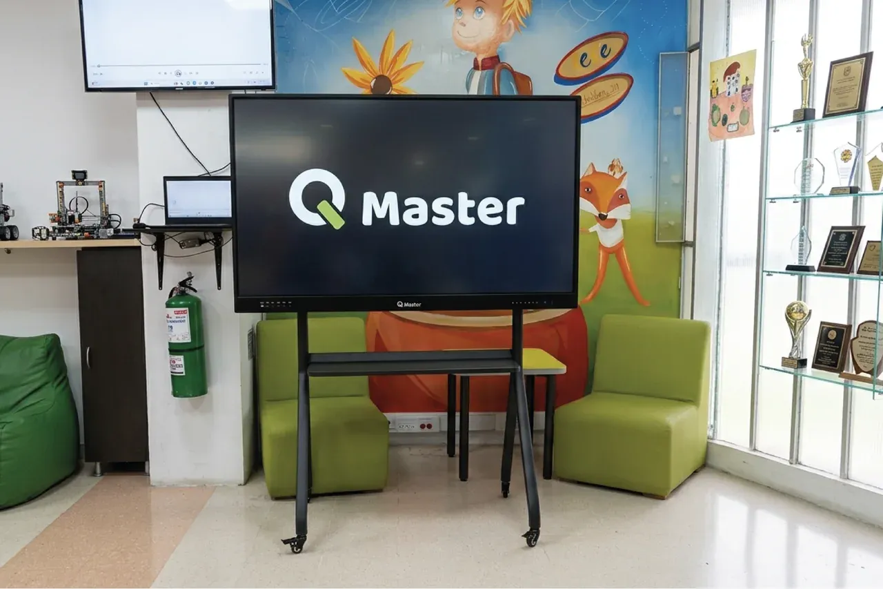Pantalla interactiva QMaster sobre soporte móvil en un aula con mobiliario infantil y decoración en la pared.