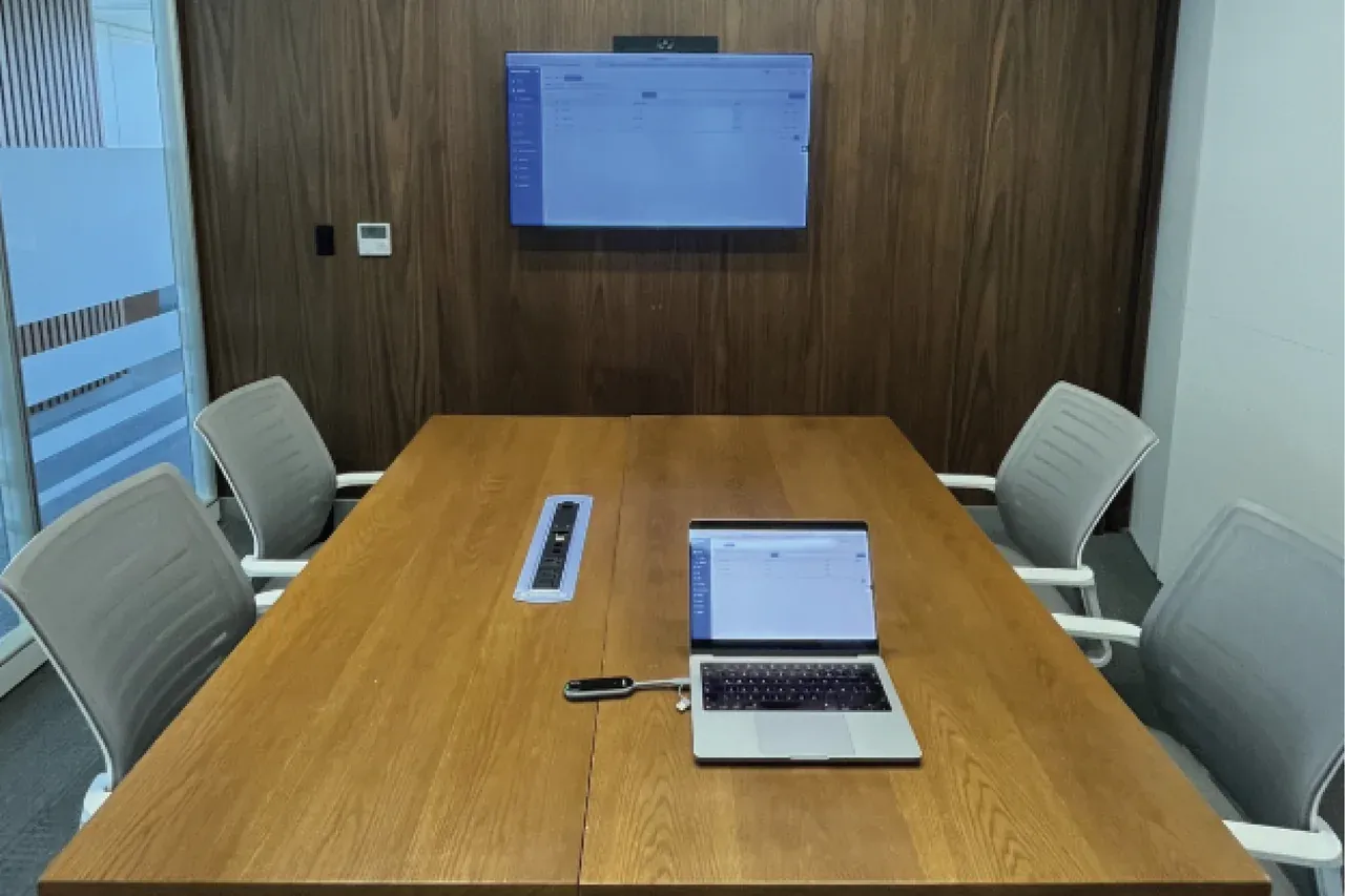 Sala de reuniones con mesa alargada, un portátil abierto sobre la madera y la pantalla encendida al fondo.
