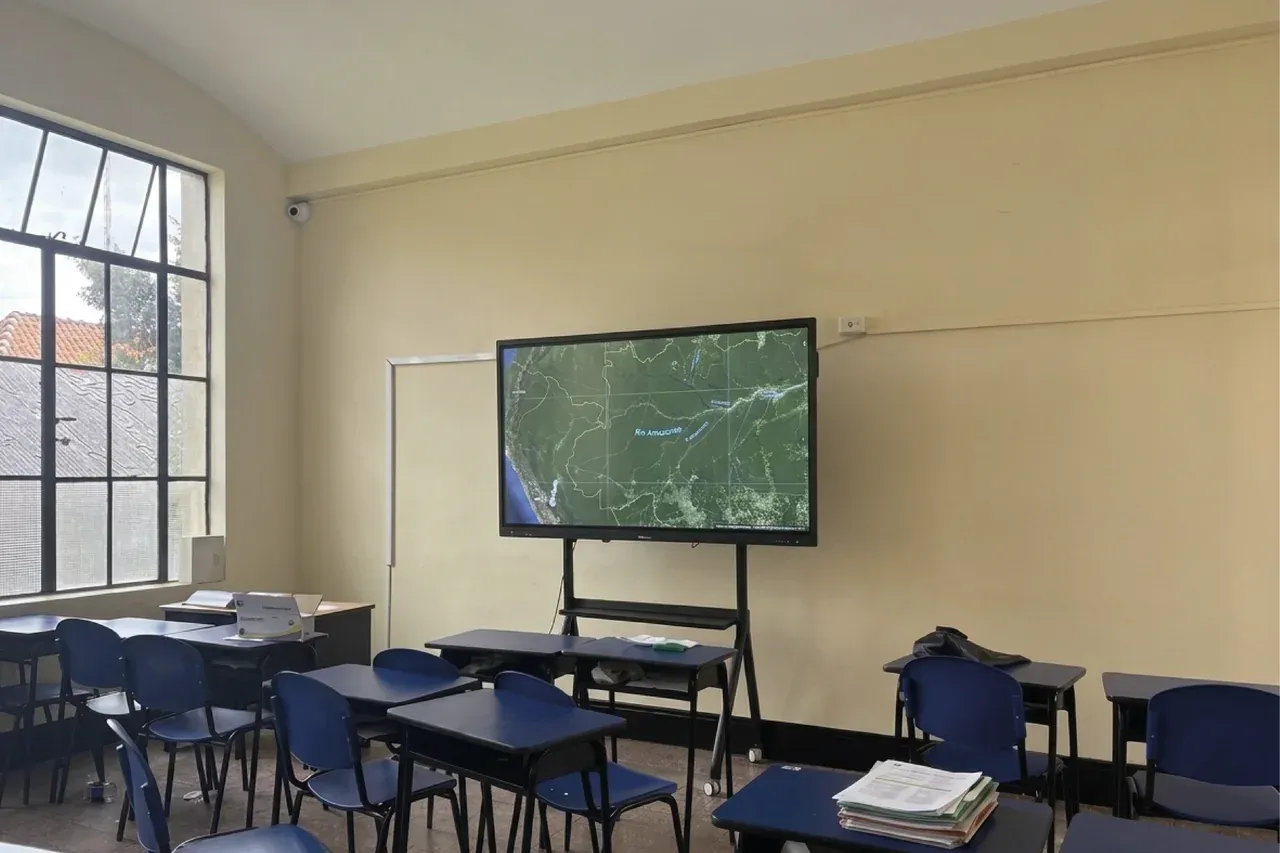 Salón con mesas de trabajo agrupadas y la pantalla interactiva QMaster encendida al frente, junto al tablero verde.