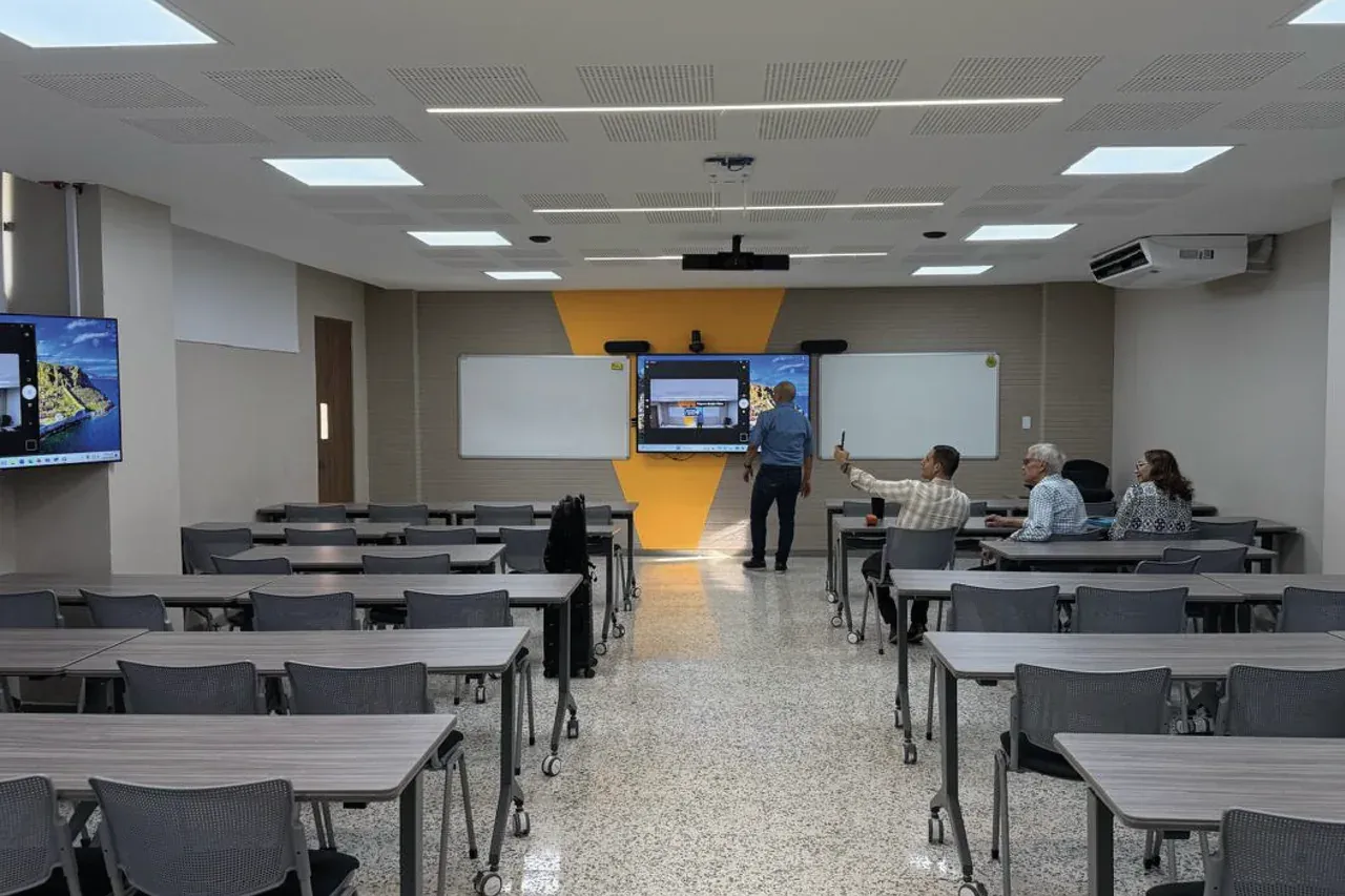 Clase híbrida en curso: estudiantes en el aula y la pantalla interactiva encendida al frente, con las cámaras montadas en el techo.