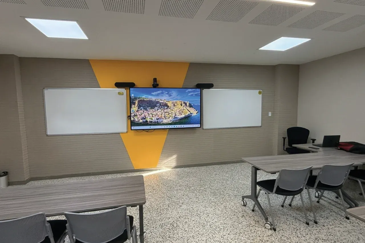 Salón con mesas móviles agrupadas y la pantalla interactiva al frente, entre dos tableros blancos.