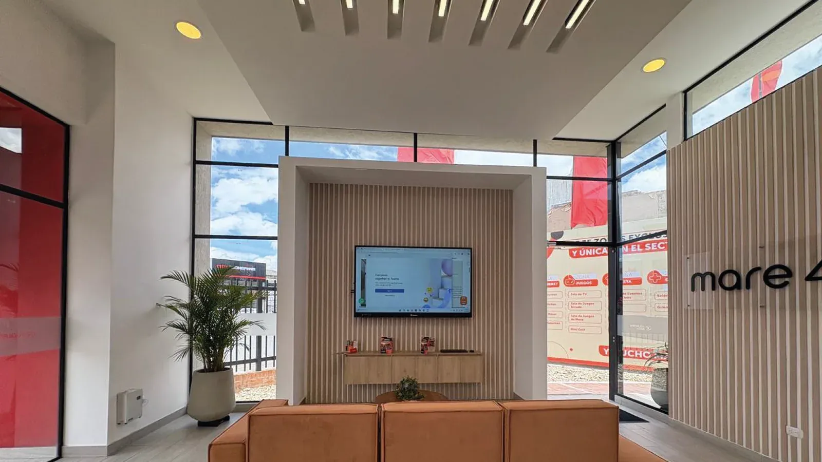 Sala de venta de Colpatria: pantalla interactiva QMaster empotrada en un nicho de madera al fondo, frente a la zona de atención con sofás y ventanales a la calle.