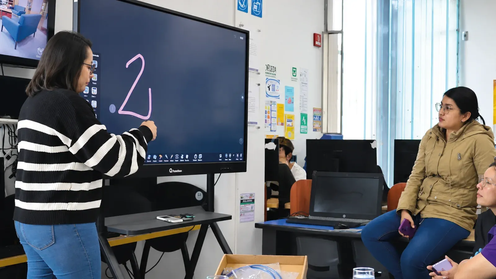 Sala de la Biblioteca Pública de Bosa: una pantalla interactiva QMaster de 65 pulgadas sobre soporte móvil, con una asistente escribiendo en la pizarra digital ante un grupo sentado.